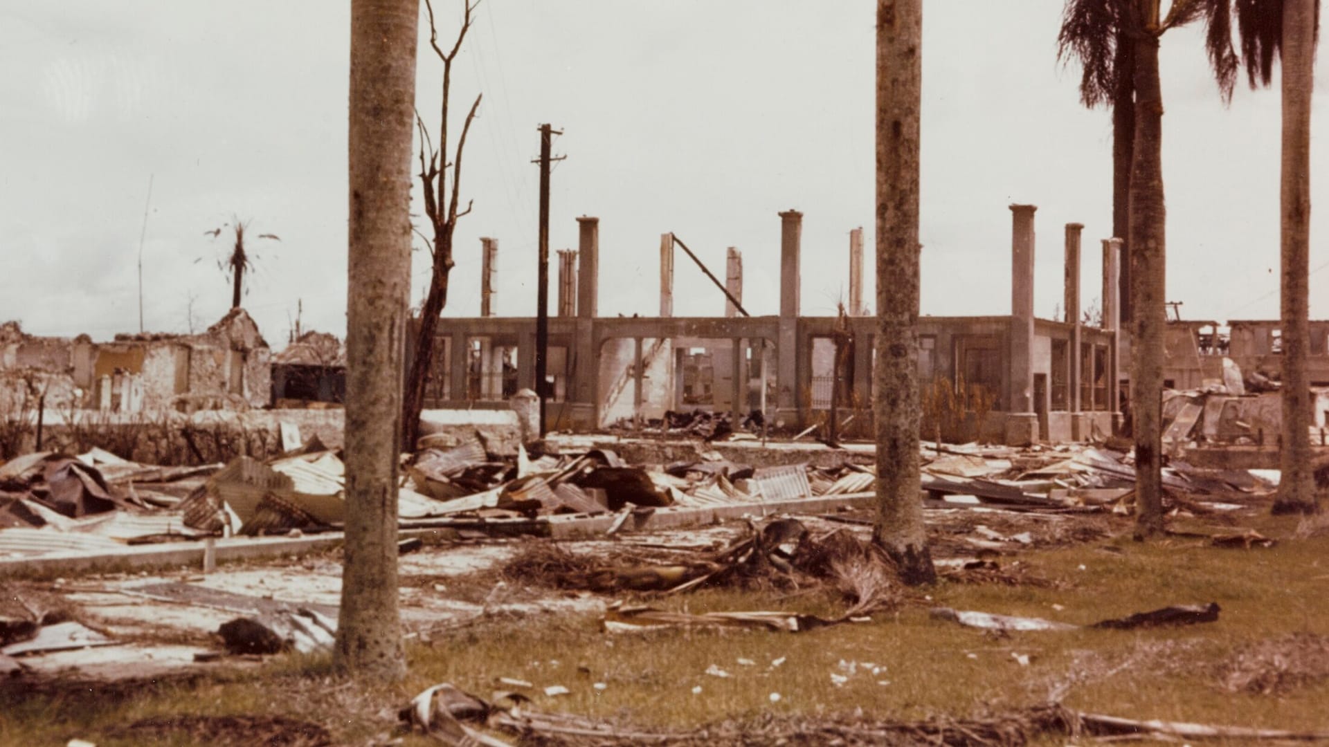 Sepia 1944 photo of a gutted old post office in Guam: roofless, only walls and columns standing amid debris, with palm trunks framing the foreground.